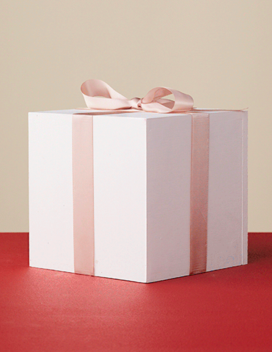 A white gift box with a pink ribbon and bow, placed on a red surface against a beige background.