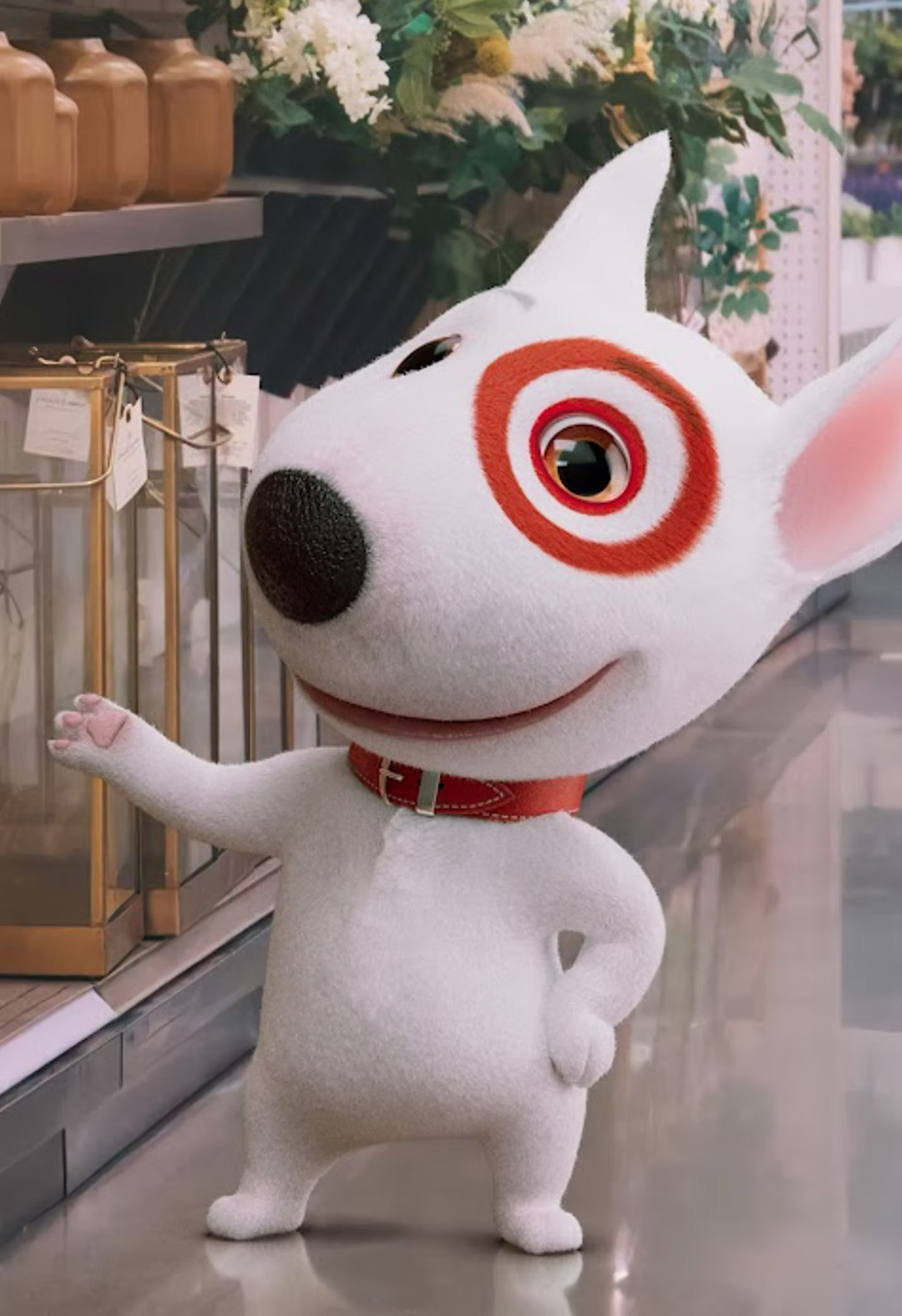 A costumed character of a white dog with a black nose, one eye surrounded by a red target design, and a red collar, standing indoors near flowers and decorative items.