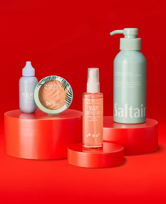 Collection of skincare and cosmetic products including a small light purple bottle, a round container of blush, a peach-colored spray bottle labeled 'Glow Baby,' and a tall mint green pump bottle labeled 'Saltair,' displayed on pink cylindrical platforms against a red background.