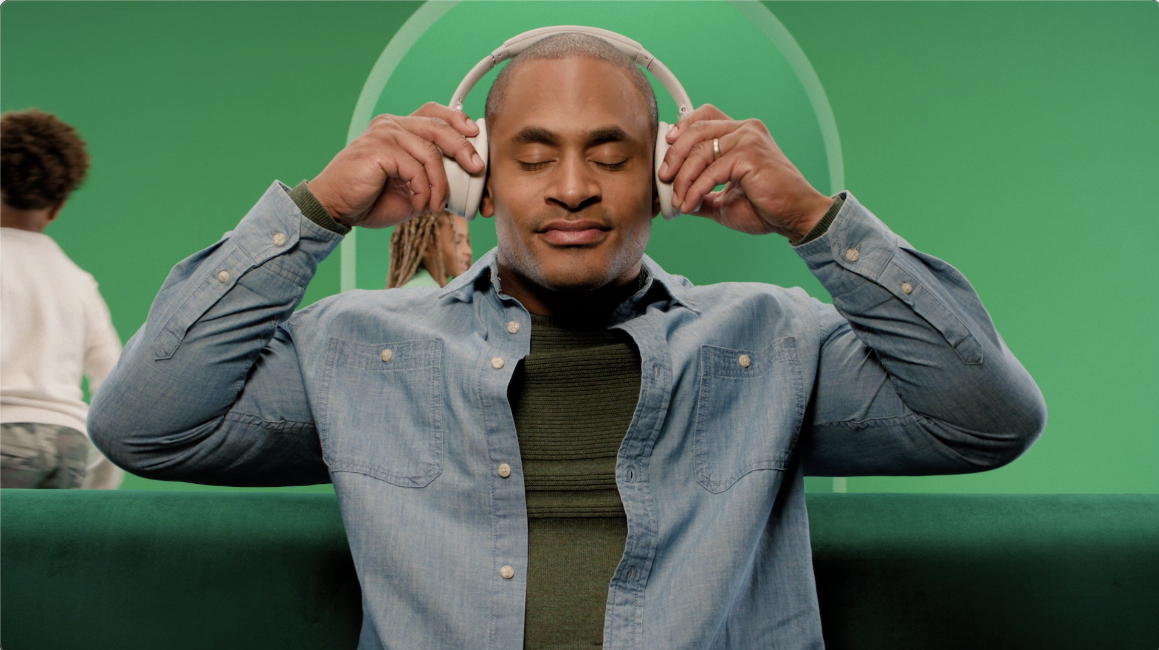 A man with closed eyes holding headphones over his ears, sitting in front of a green screen. Two other people are in the background.