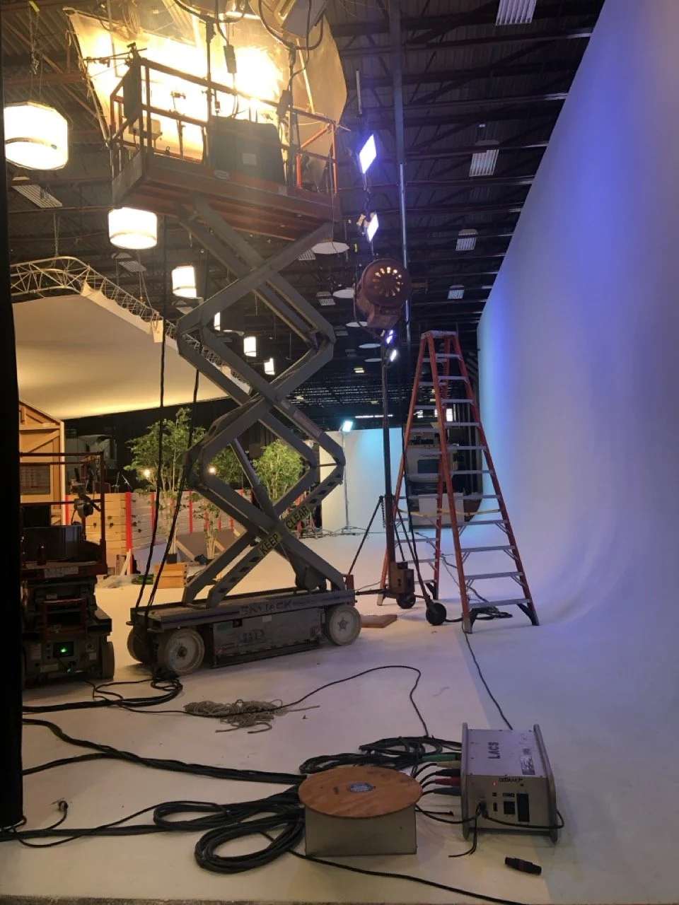 A photo of a studio set under construction with lighting equipment, a blue backdrop, a ladder, a scissor lift, and various cables and electrical equipment.