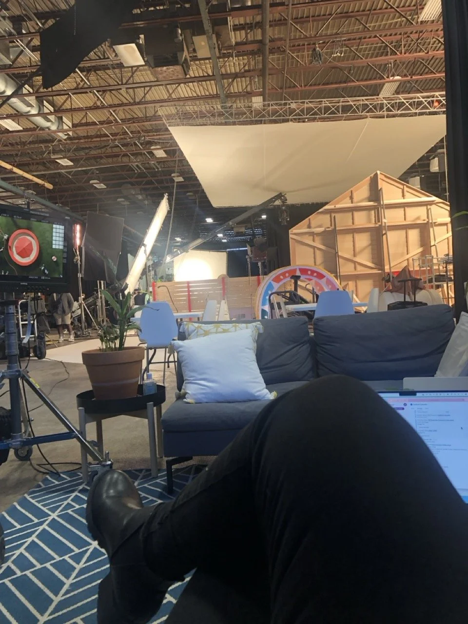 View from someone sitting on a rug with crossed legs, shows a studio or film set with lighting equipment, a potted plant, a sofa with pillows, and various set pieces including a large circular prop and wooden structures, with a high industrial ceiling.