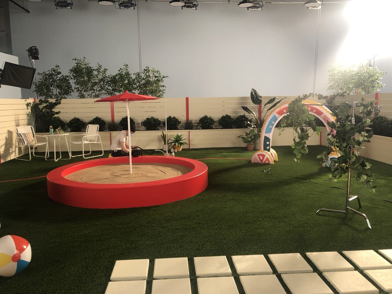 Indoor space decorated with a red sandbox, a small rainbow arch, a beach ball, potted plants, white chairs, a table, and an umbrella on artificial grass.