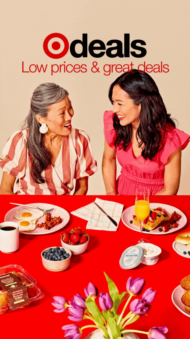 Two women smiling at each other with a breakfast spread on a red table, including eggs, bacon, dessert, berries, fruit, yogurt, and flowers, with a Target logo and the words "deals Low prices & great deals" above them.