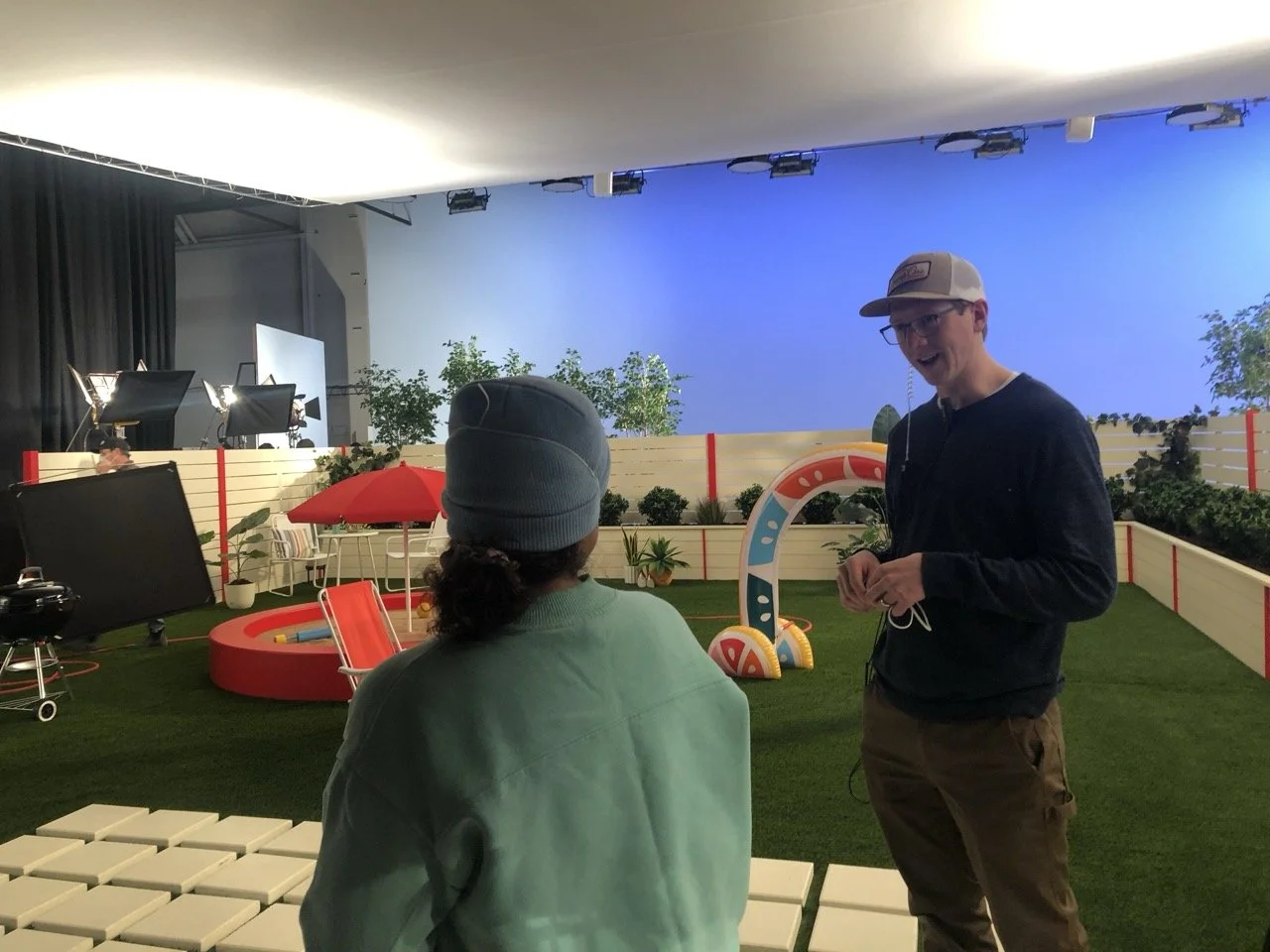 A behind-the-scenes shot of a studio set with a small outdoor scene, including a red slide, a red umbrella, plants, and a blue background, with two men talking and filming equipment visible.