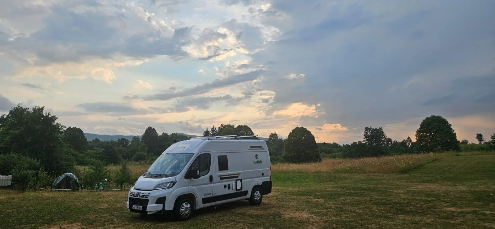A white camper van parked in a grassy field with trees and a cloudy sky in the background.