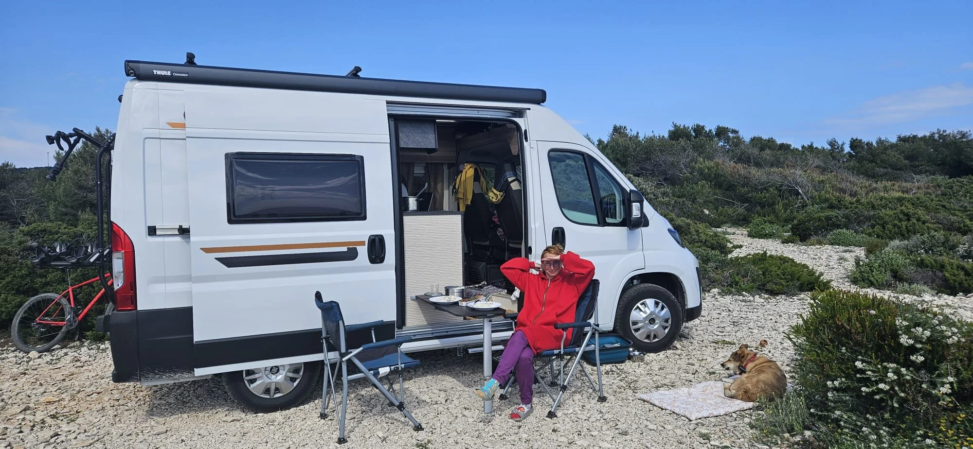 A woman in a red jacket sitting outside a white camper van with a dog resting on a blanket nearby, in a rocky outdoor setting under a blue sky.