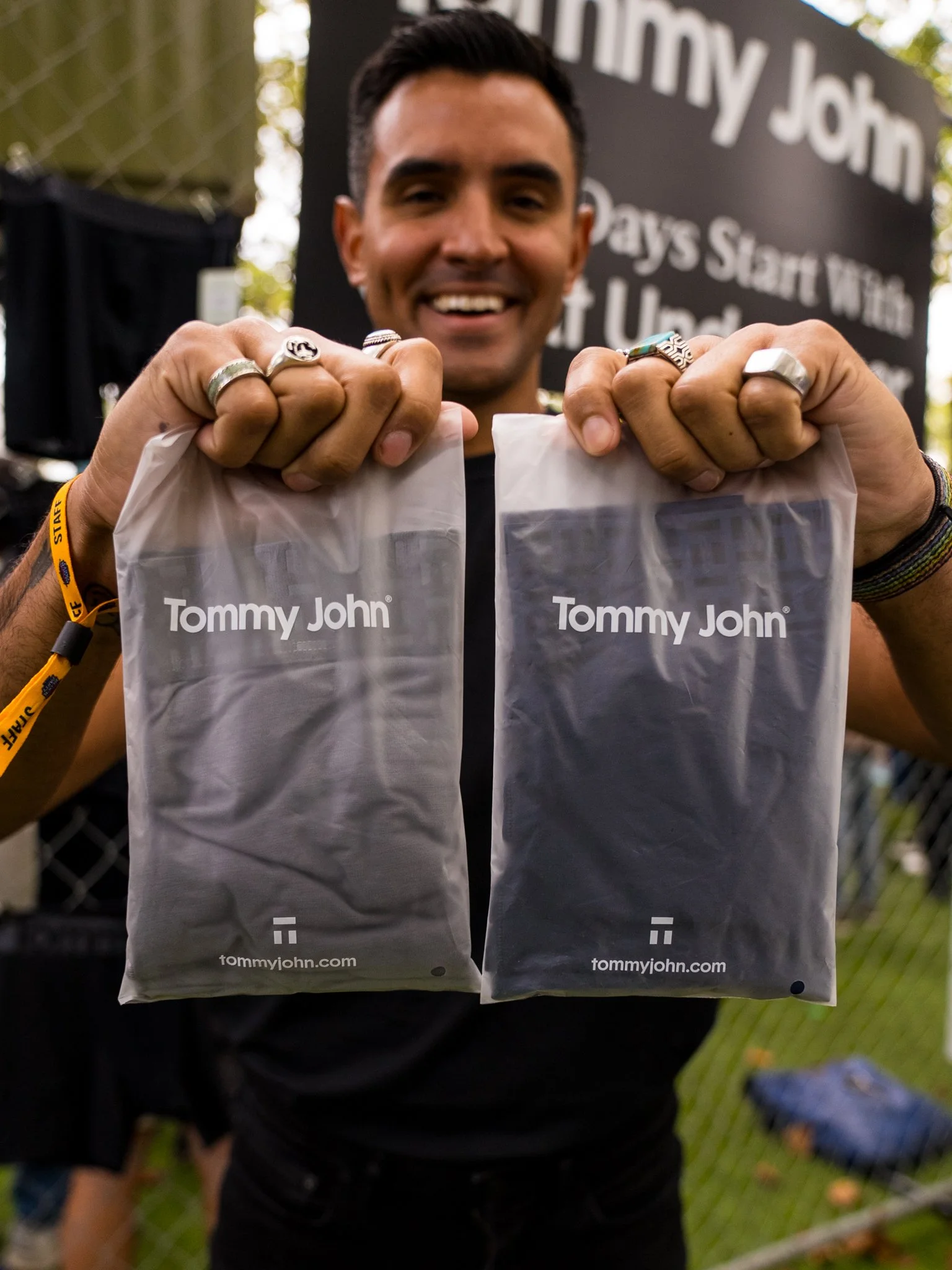 A man smiling and holding two clear plastic bags with 'Tommy John' written on them, outdoors in front of a sign that says 'Tommy John.'