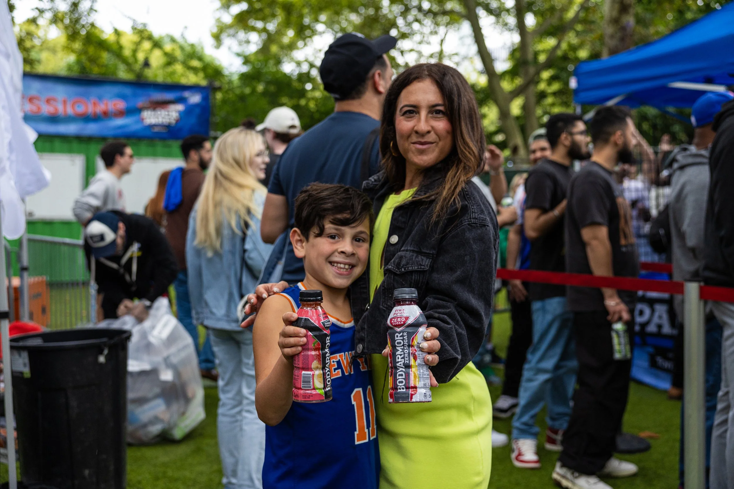 A woman and a young boy holding bottles of Bodyarmor sports drinks at an outdoor event with a crowd of people and trees in the background.