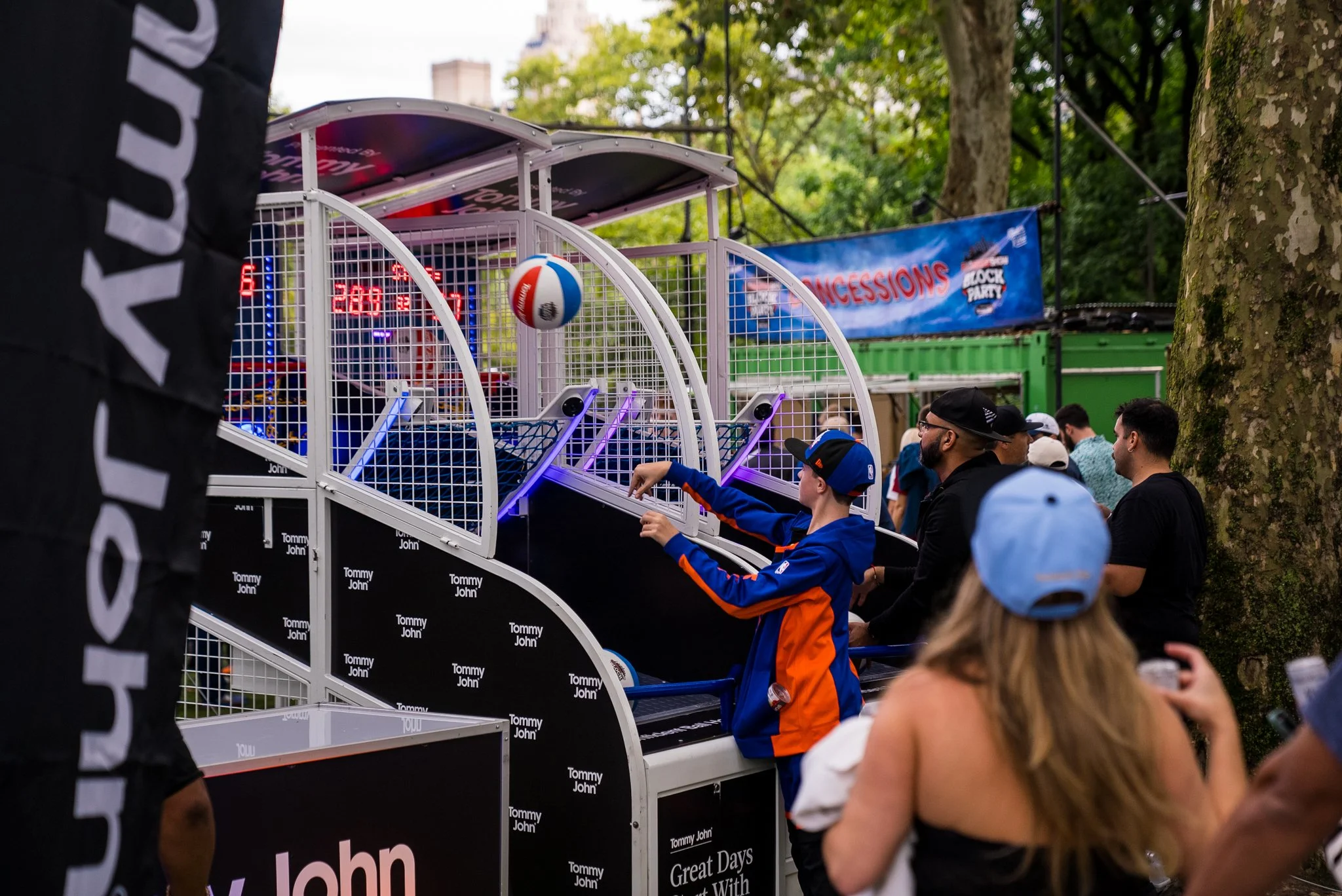 People playing a ball game at an outdoor carnival or fair with trees and banners in the background.