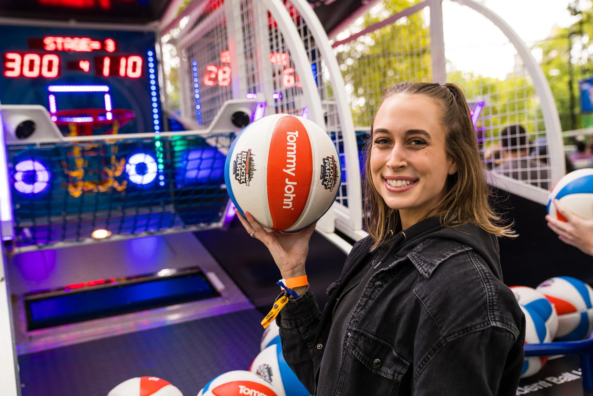A young woman smiling and holding a colorful volleyball with her right hand at an arcade basketball game. The game features digital score displays and several other volleyballs on the ground nearby.