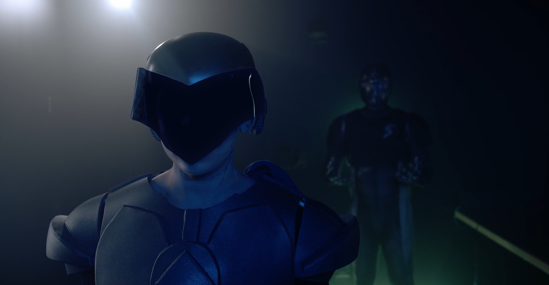 Characters in masks facing the camera, one behind the other, with a blue hue