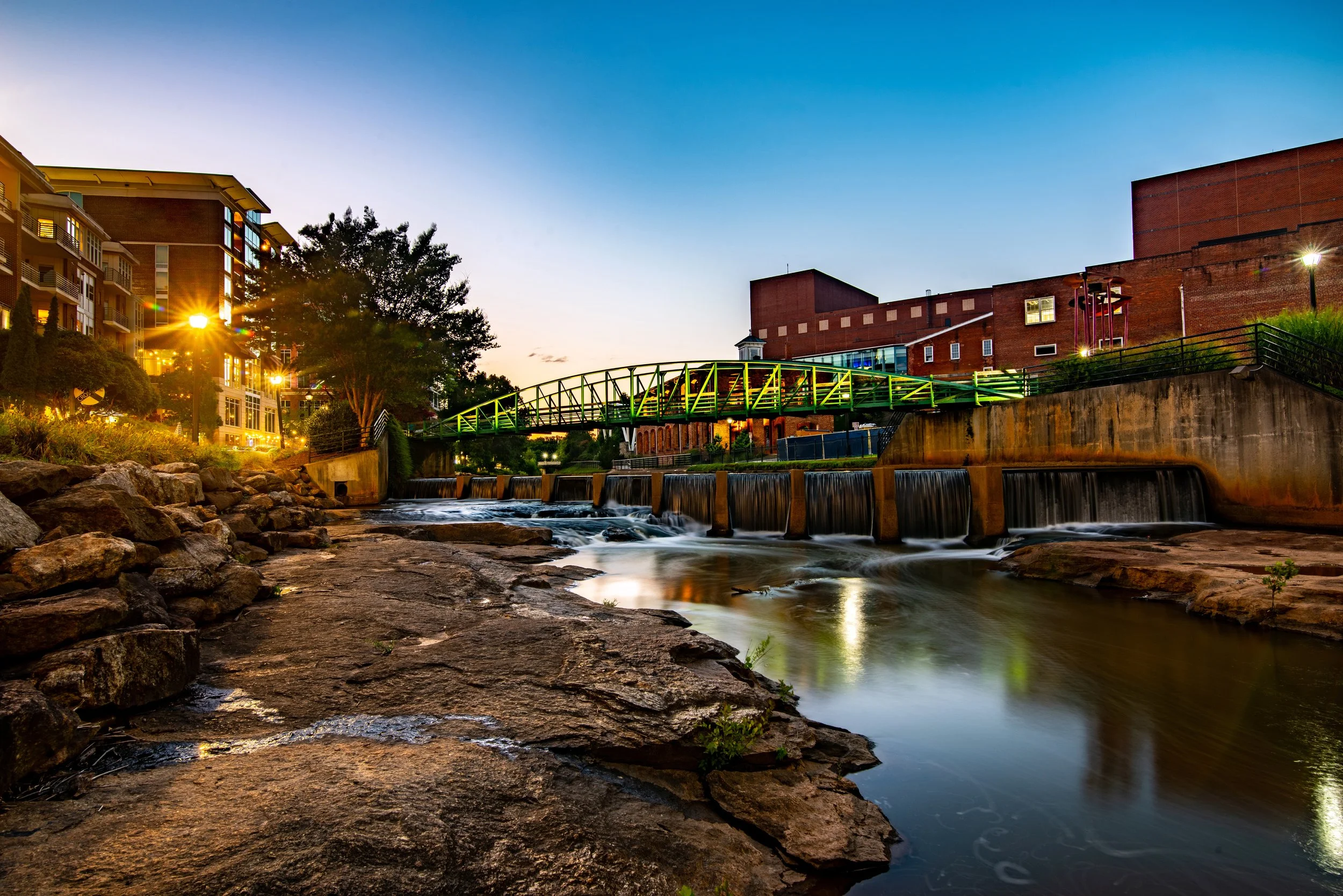 Outdoor scene of a creek with small waterfalls, a bridge with green railings, and buildings in the background at sunset. Reedy River in Greenville, South Carolina.