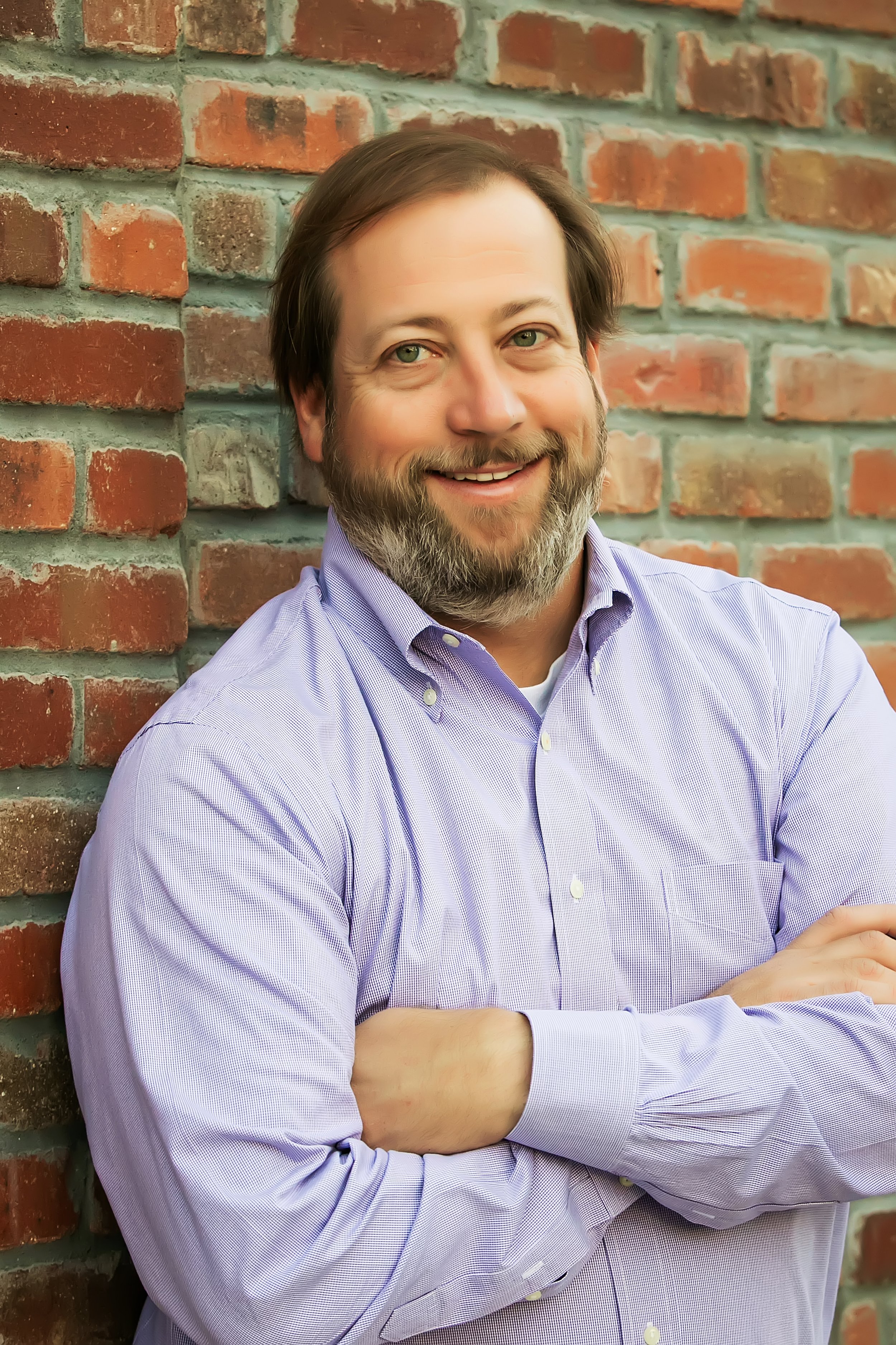 A smiling man with a beard and light brown hair, wearing a dark blazer and a light-colored shirt, sitting indoors.