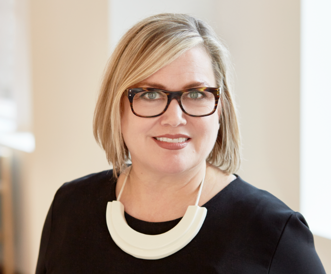 A woman with blonde hair, wearing glasses, a black top, and a white statement necklace, smiling indoors.