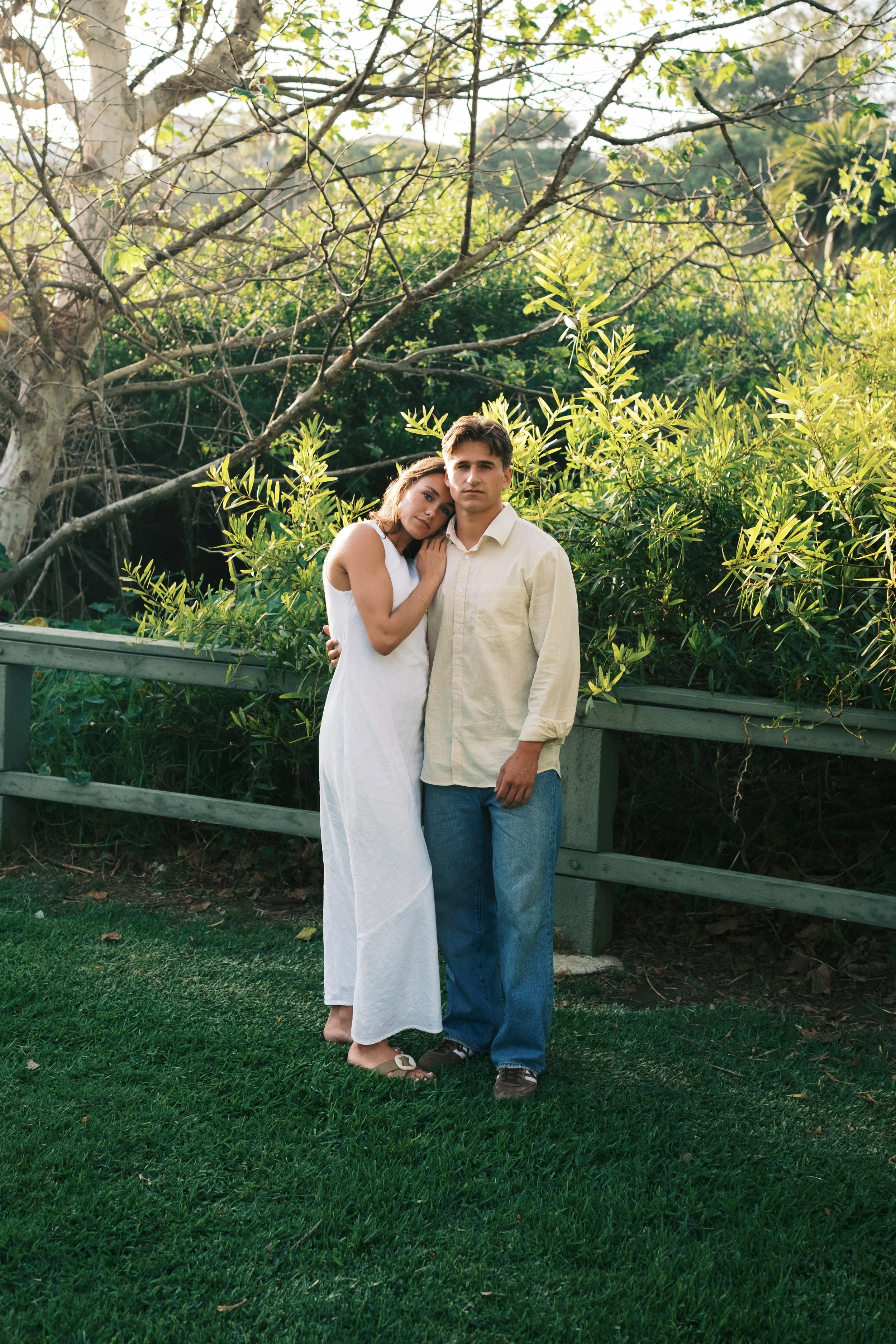 A young couple stands close together outdoors among green foliage and trees, with a wooden railing nearby. The woman leans her head on the man's shoulder and they look directly at the camera, both dressed in light-colored clothing.