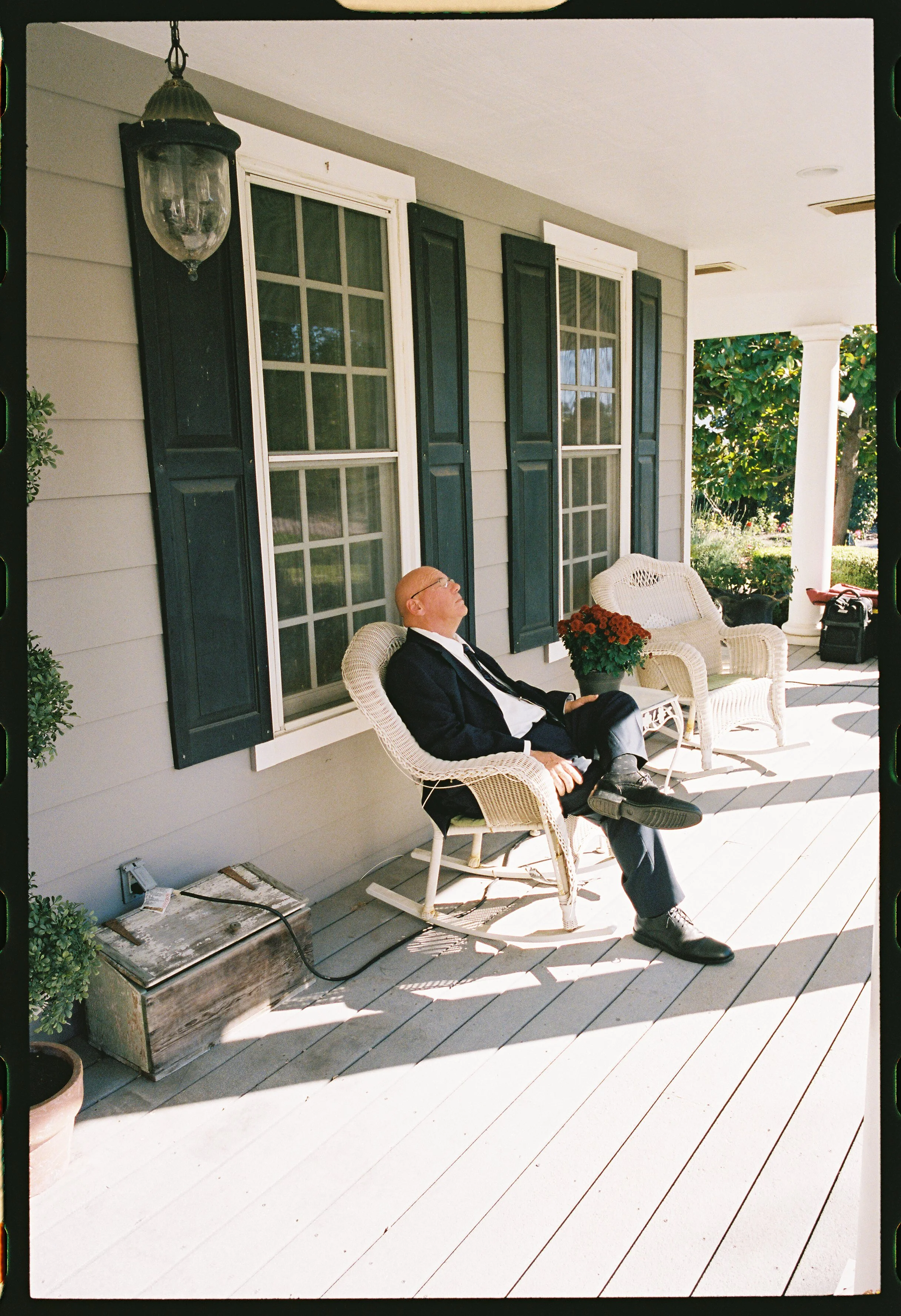 A man in a tuxedo is sitting on a porch in a wicker rocking chair, relaxing with his eyes closed, wearing glasses. There is a potted red flower next to him on the porch, which has white wooden flooring, black window shutters, and a grey house exterio