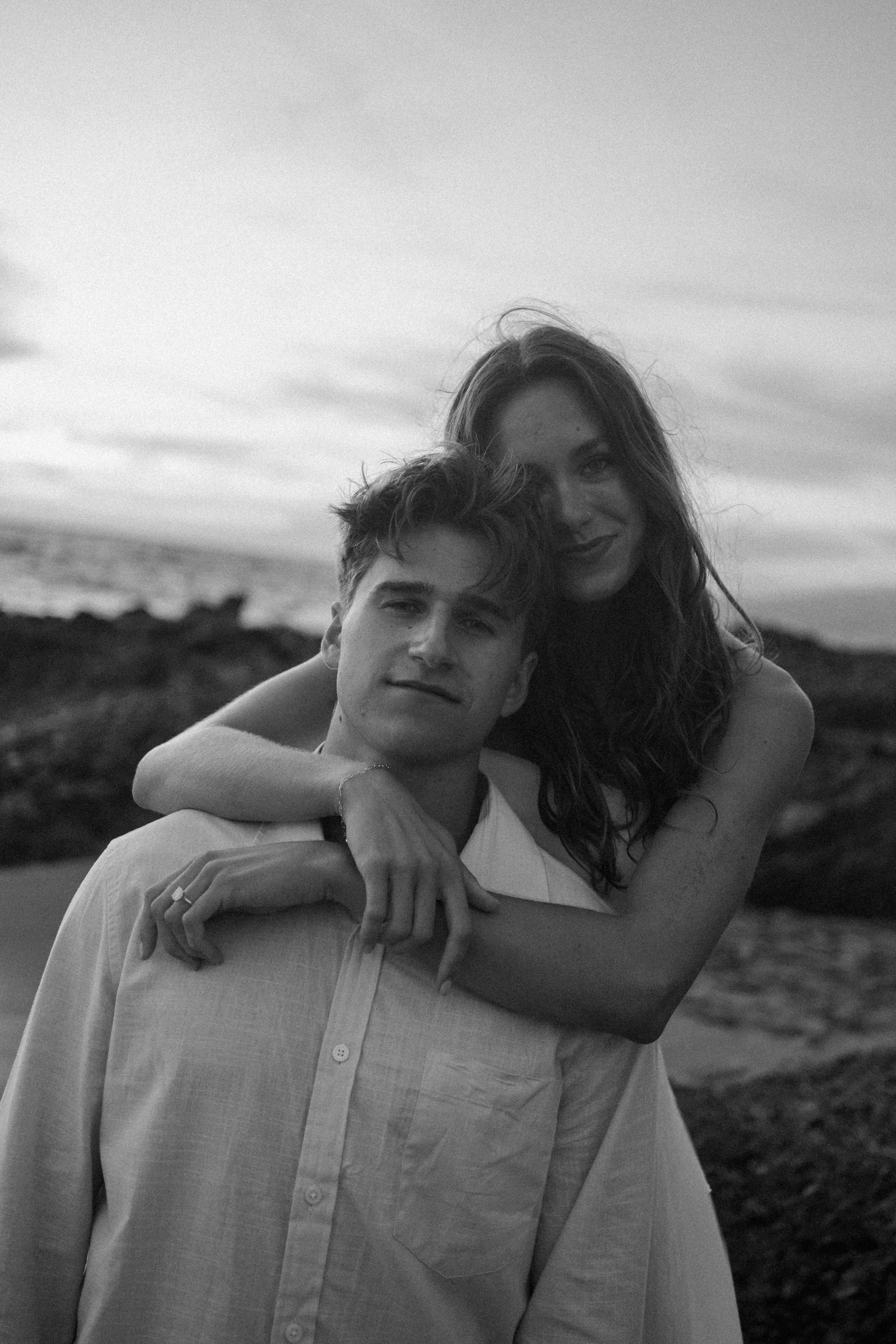 A young man and a woman are hugging outdoors near the beach during sunset or sunrise, in black and white.