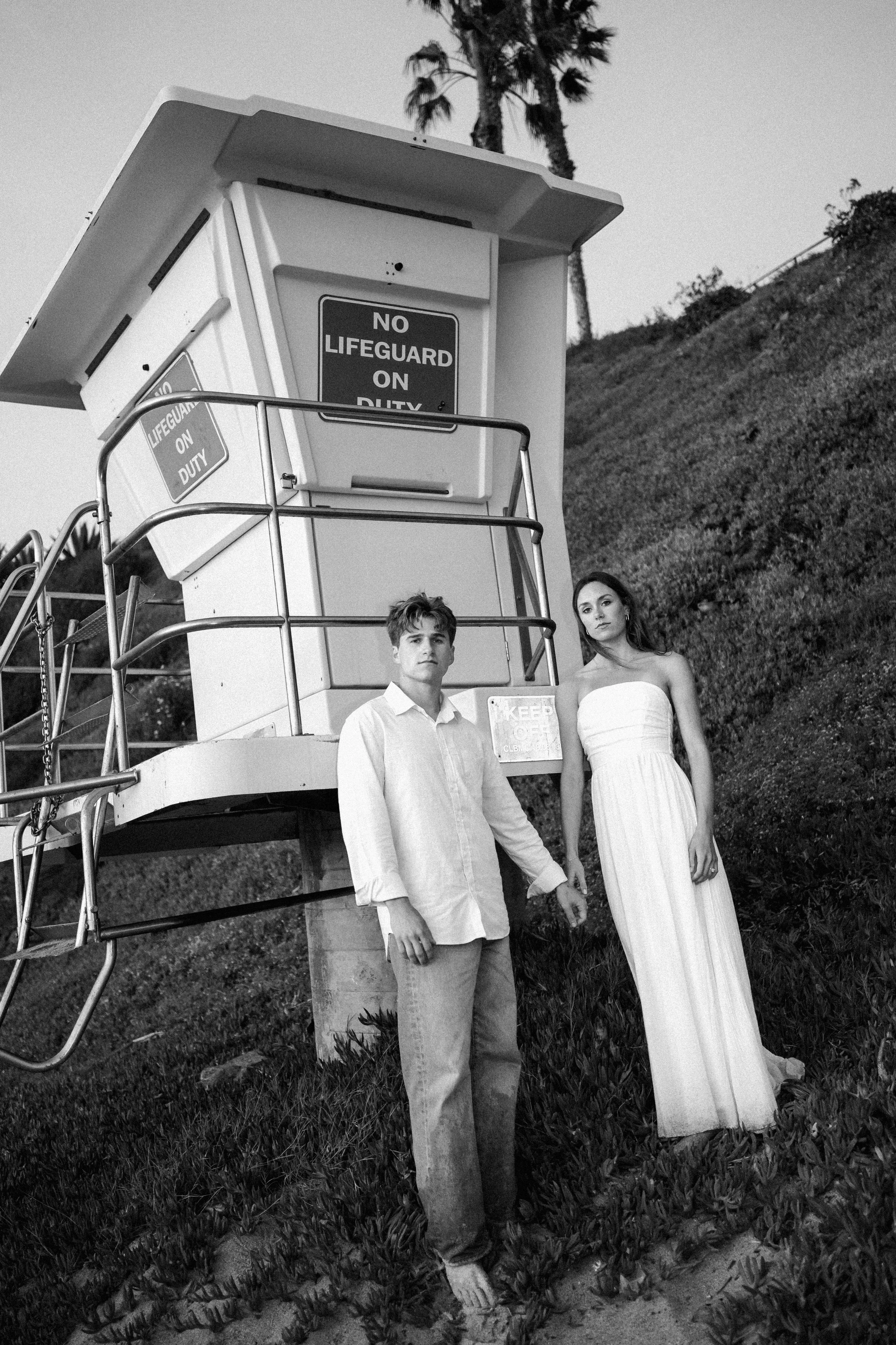 Black and white photo of a man and woman standing beside a lifeguard tower at the beach, holding hands. The man is wearing a button-up shirt and pants, the woman is in a strapless dress. Palm trees are visible in the background.