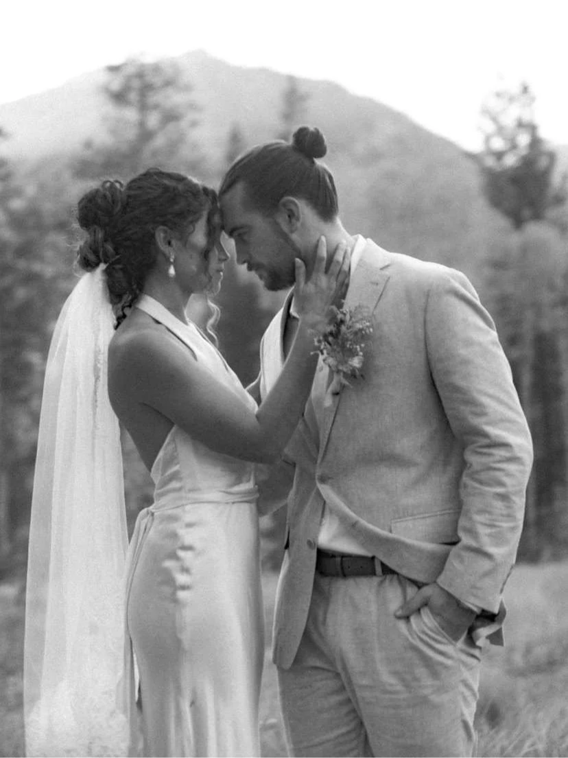 Black and white photo of a bride and groom with foreheads touching, outdoors with trees and mountains in the background. The bride is young, wearing a satin wedding dress with a veil, and has curly hair. The groom is young, wearing a light-colored suit and has his hair in a bun. The bride's hand is gently on the groom's face.