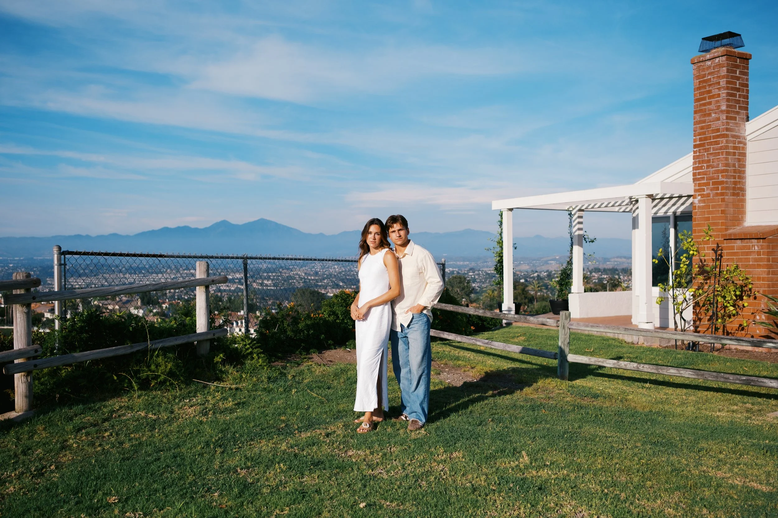 A young couple stands together on a grassy outdoor area with a city view and mountainous landscape in the background. The woman wears a white dress, and the man wears a cream shirt and blue jeans. A brick chimney and a small patio are visible near a 