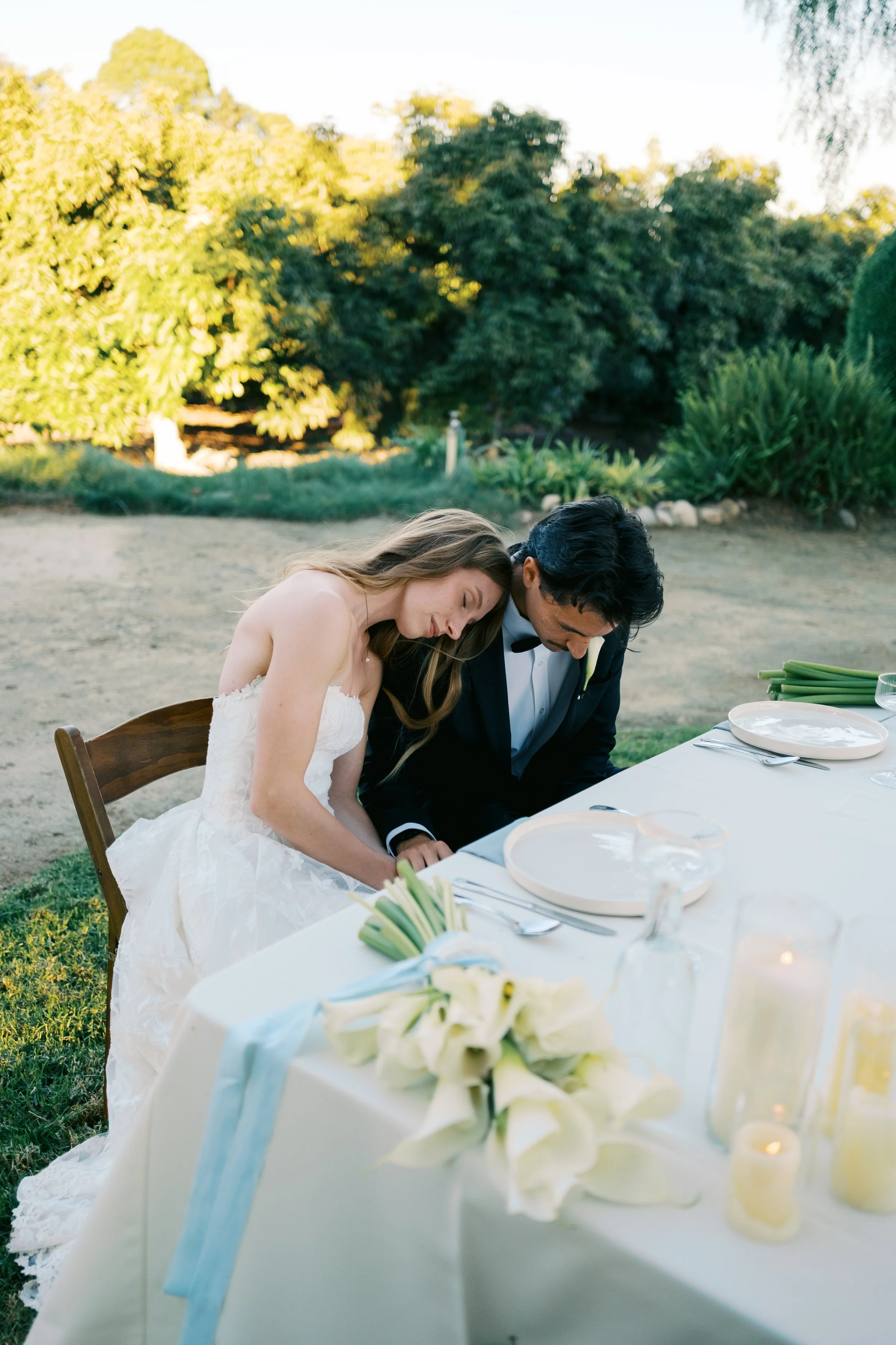 A bride and groom are sitting at a decorated outdoor wedding table, leaning their heads together. The table has white flowers, candles, plates, and cutlery, set outdoors with green trees and bushes in the background.