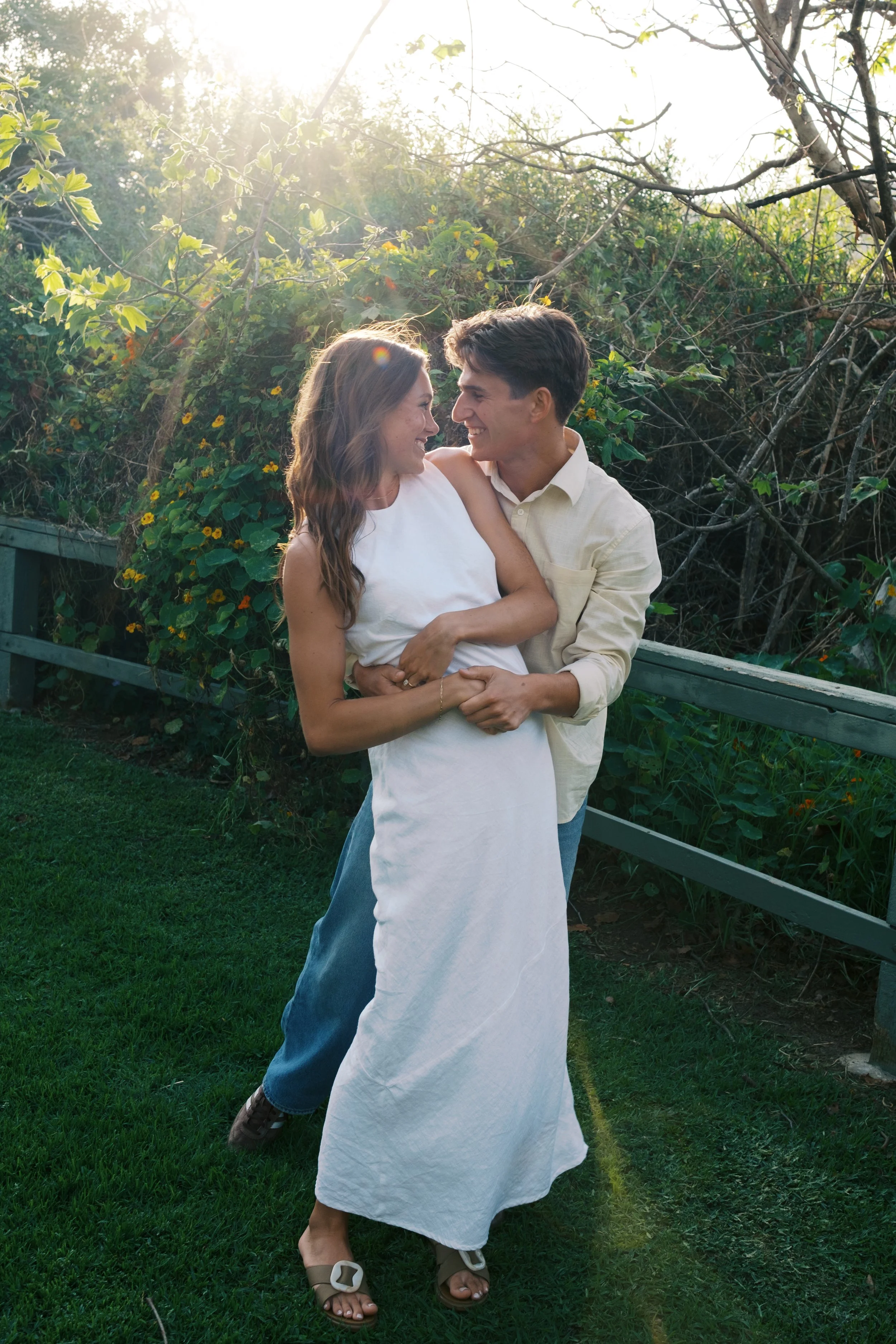 A smiling couple dancing outdoors on green grass with sunlight filtering through trees and bushes in the background.