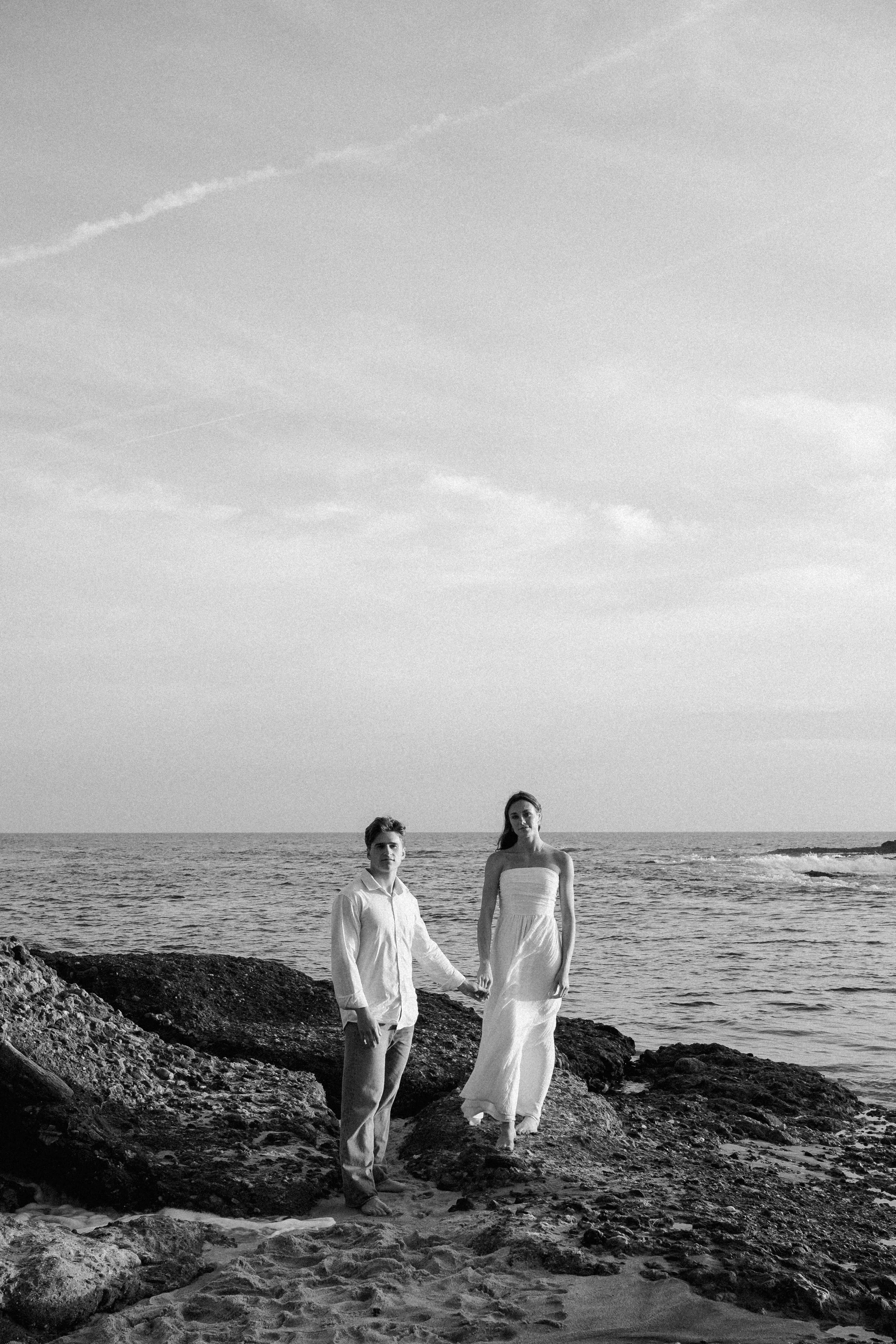 A black and white photograph of a man and woman holding hands on a rocky beach. The man is wearing a light-colored shirt and pants, and the woman is wearing a long, strapless dress. They are standing near the water's edge, with the ocean and a cloudy