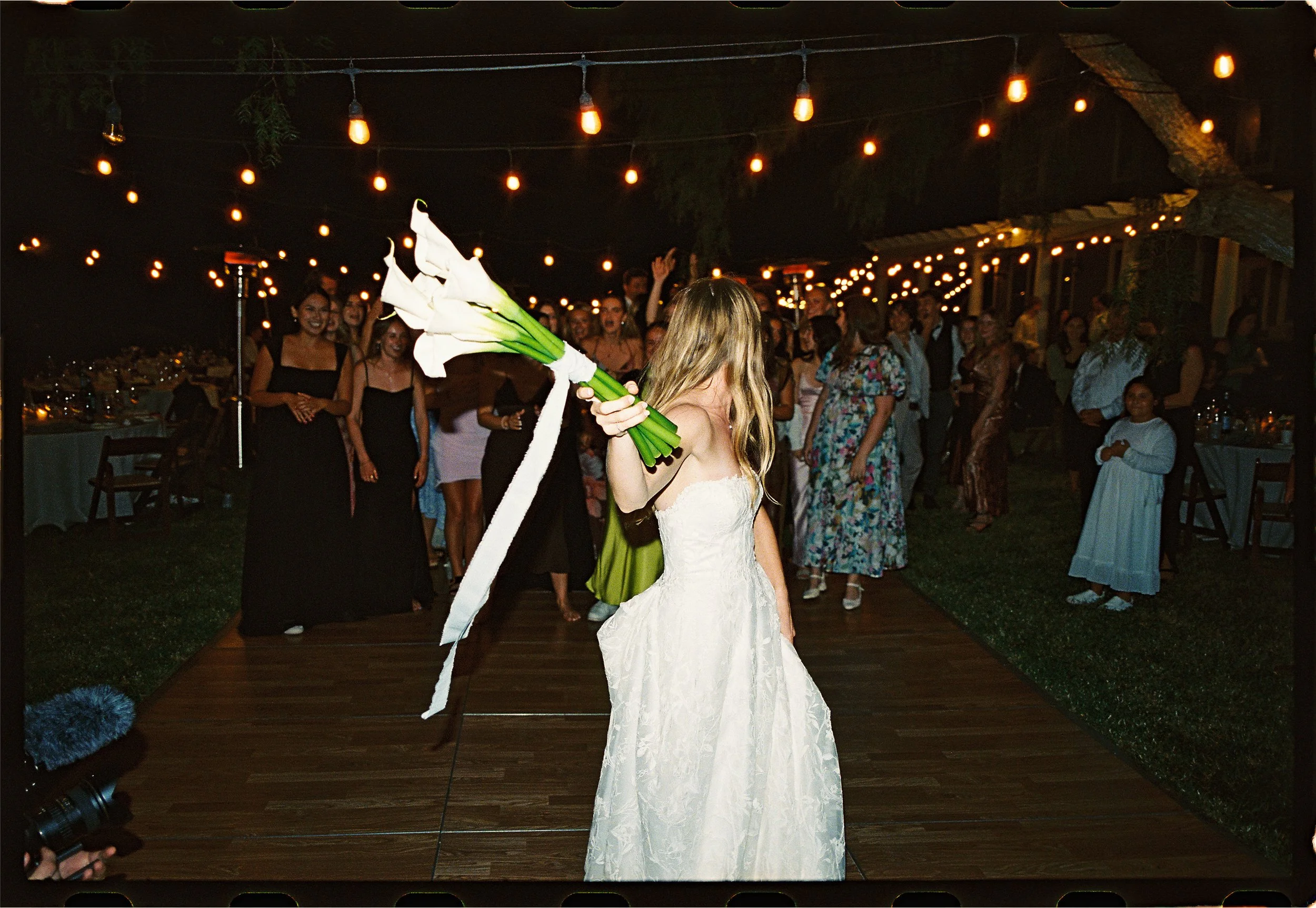 A bride in a white wedding dress holding a bouquet of white calla lilies is on a wooden dance floor at night, with guests watching and celebrating under string lights.