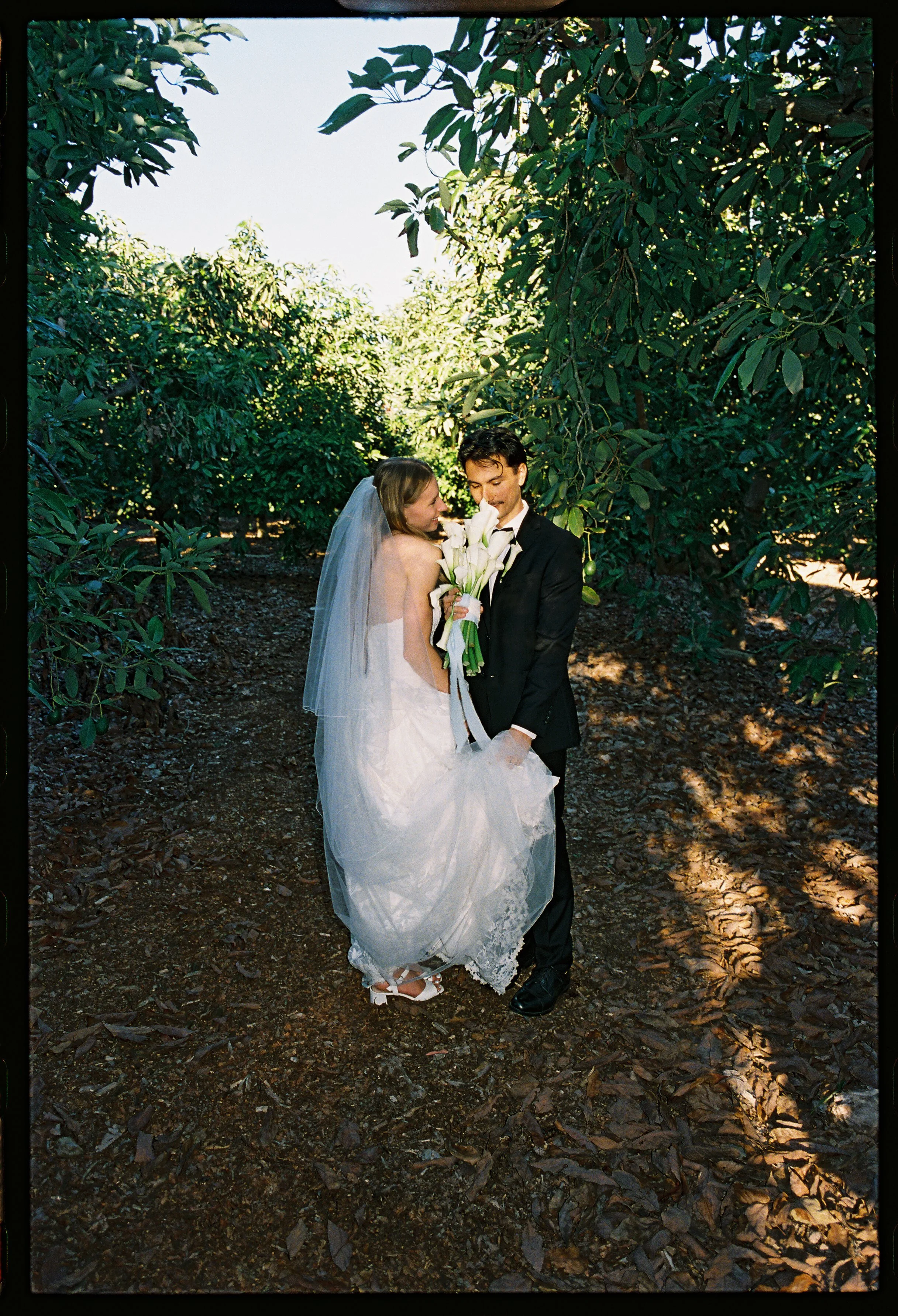 A bride and groom standing close together in a wooded area, with the bride holding a bouquet of white calla lilies and wearing a wedding dress with veil, and the groom dressed in a black suit and tie.