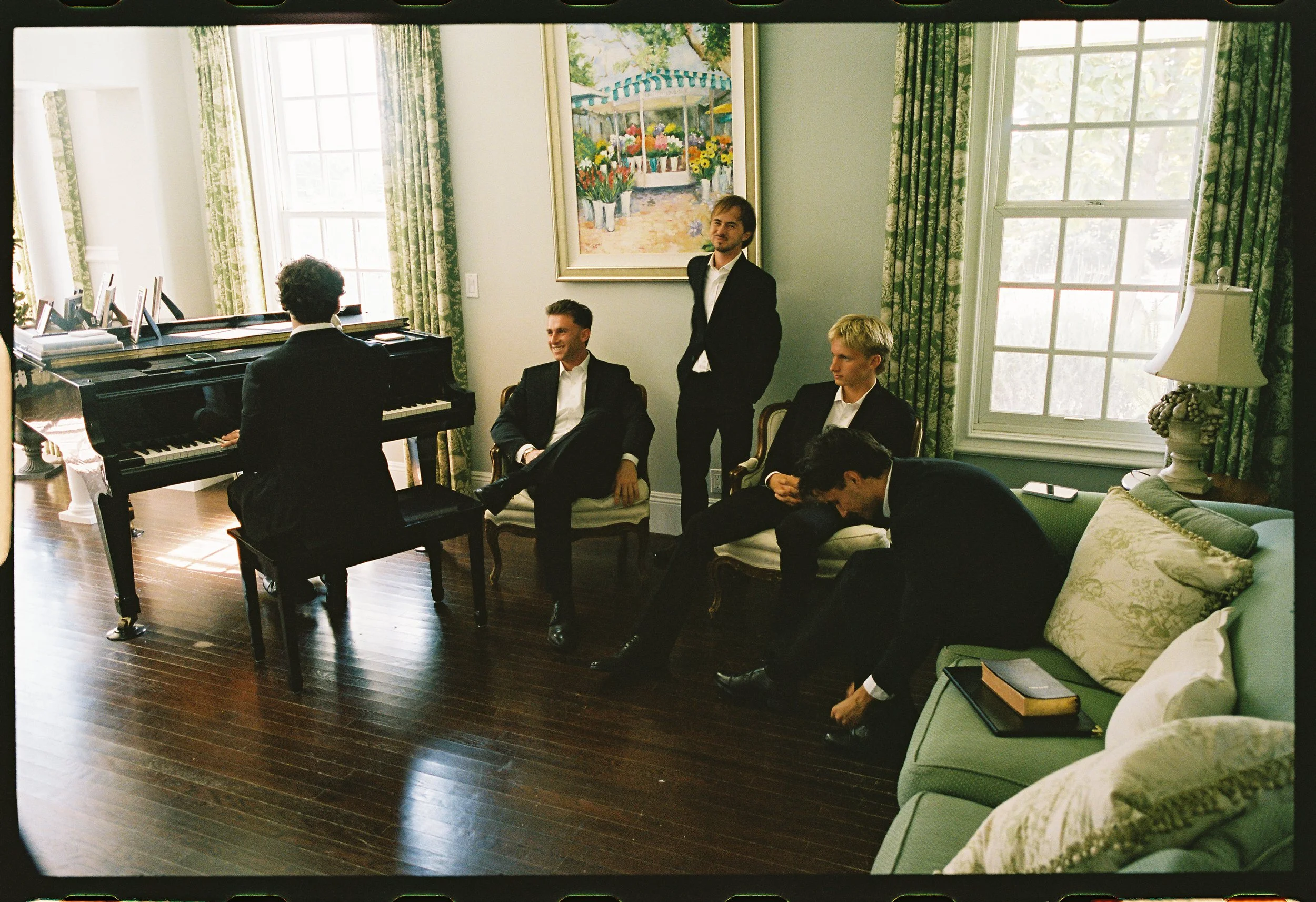 Five men in formal attire in a well-lit sitting room with a grand piano, green curtains, and framed artwork on the wall. One man is seated at the piano, four others are seated or standing nearby, engaged in conversation or adjusting shoes.