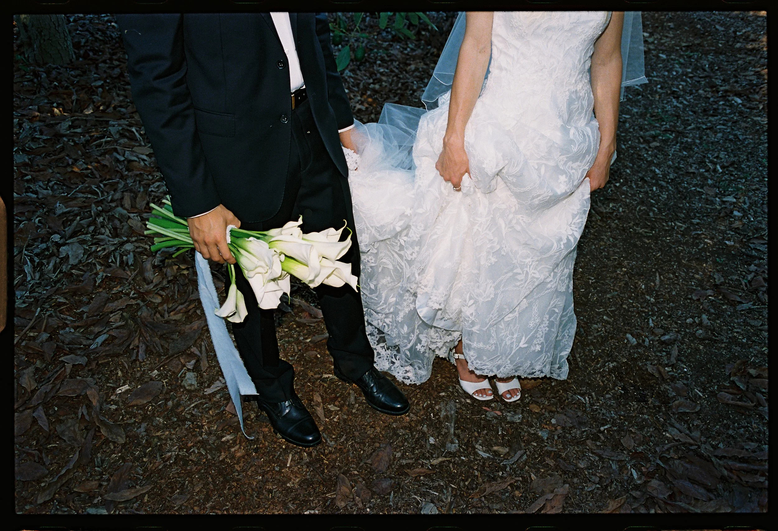 Bride in a white lace dress and heels holding her wedding dress, standing next to a groom in a black suit holding white lilies, on a dark outdoor ground.