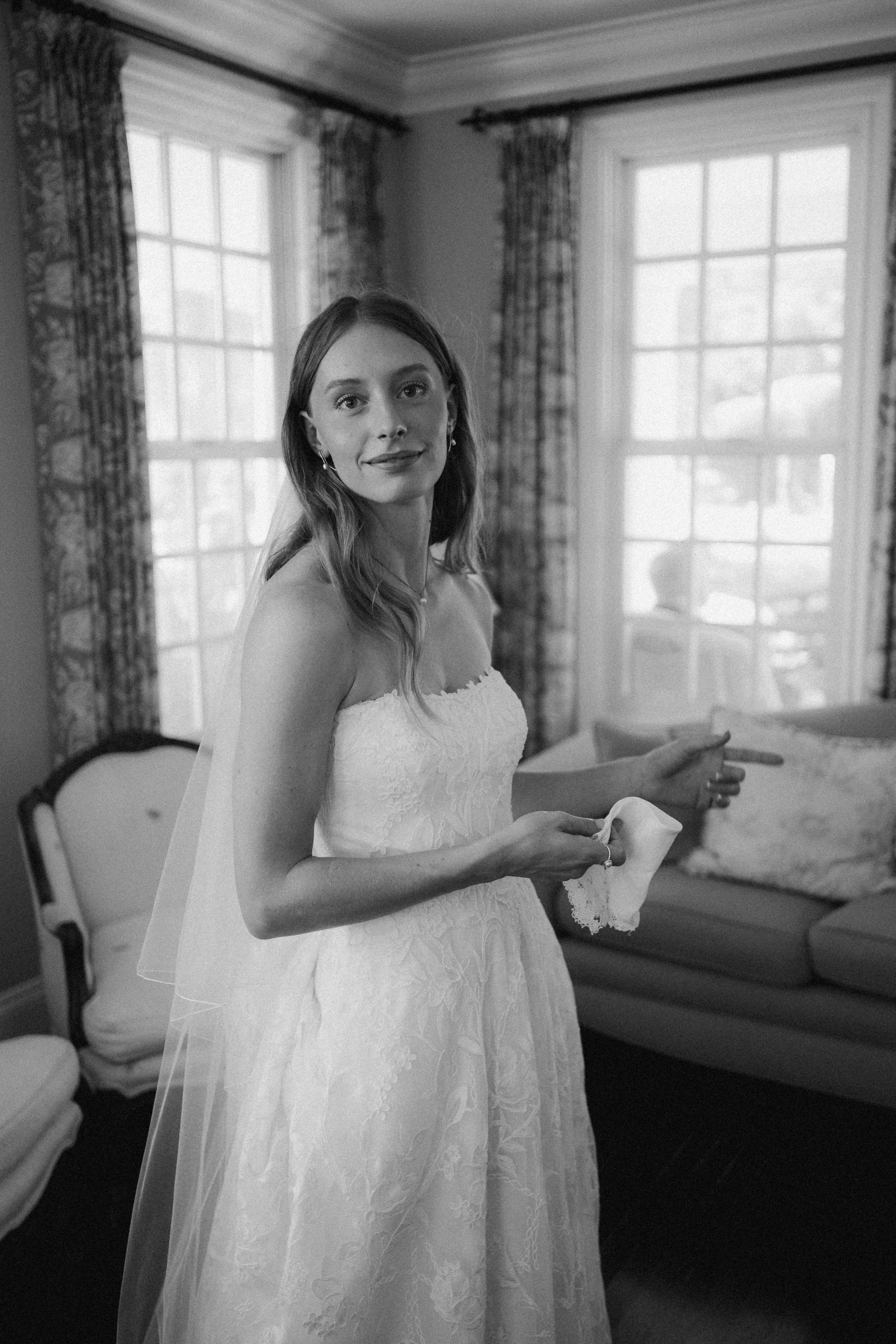 A woman in a wedding dress holding a tissue, standing indoors near windows with patterned curtains, with a sofa and armchair in the background.