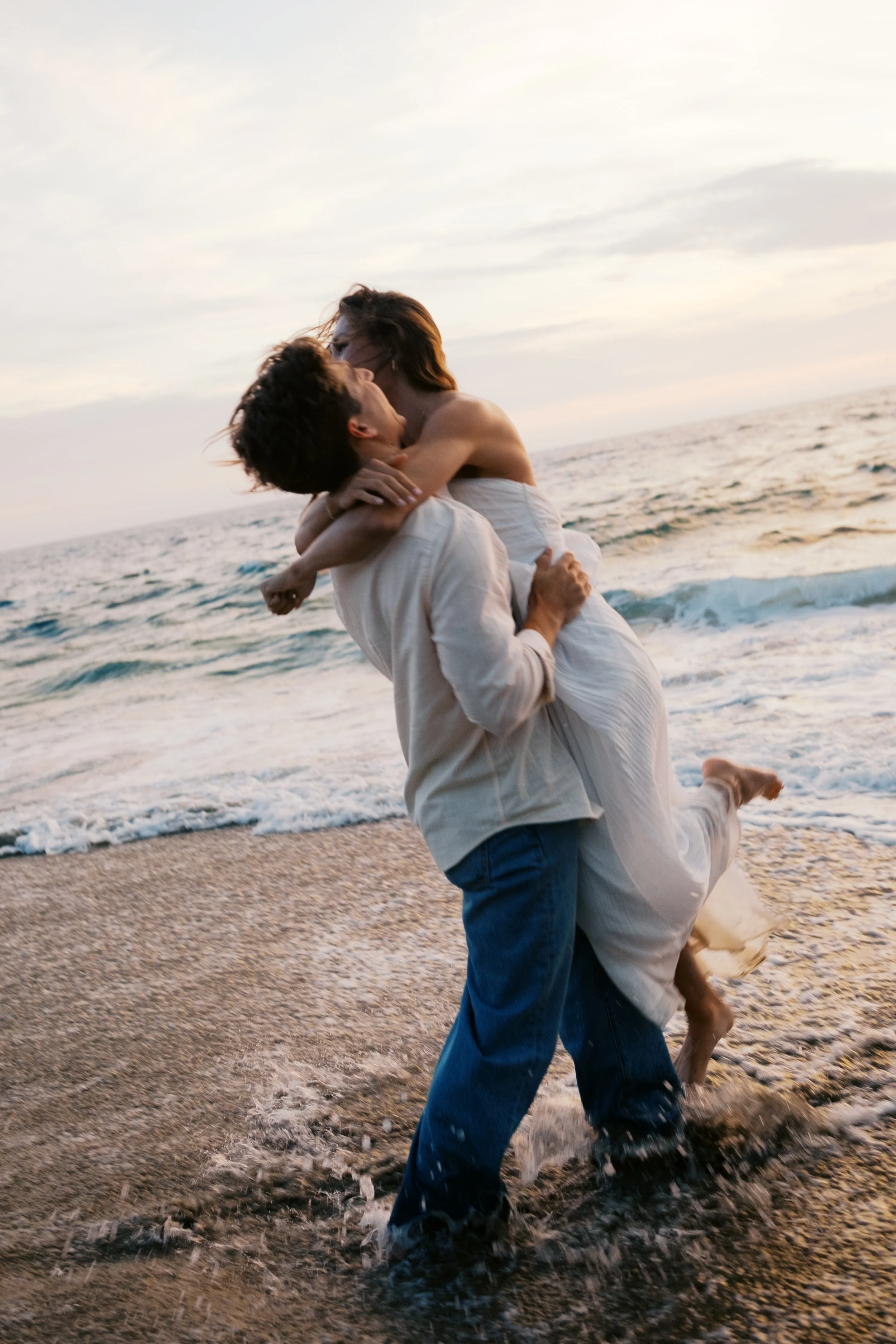 A couple is embracing and kissing on the beach with the ocean and sunset in the background, with one person lifting the other.
