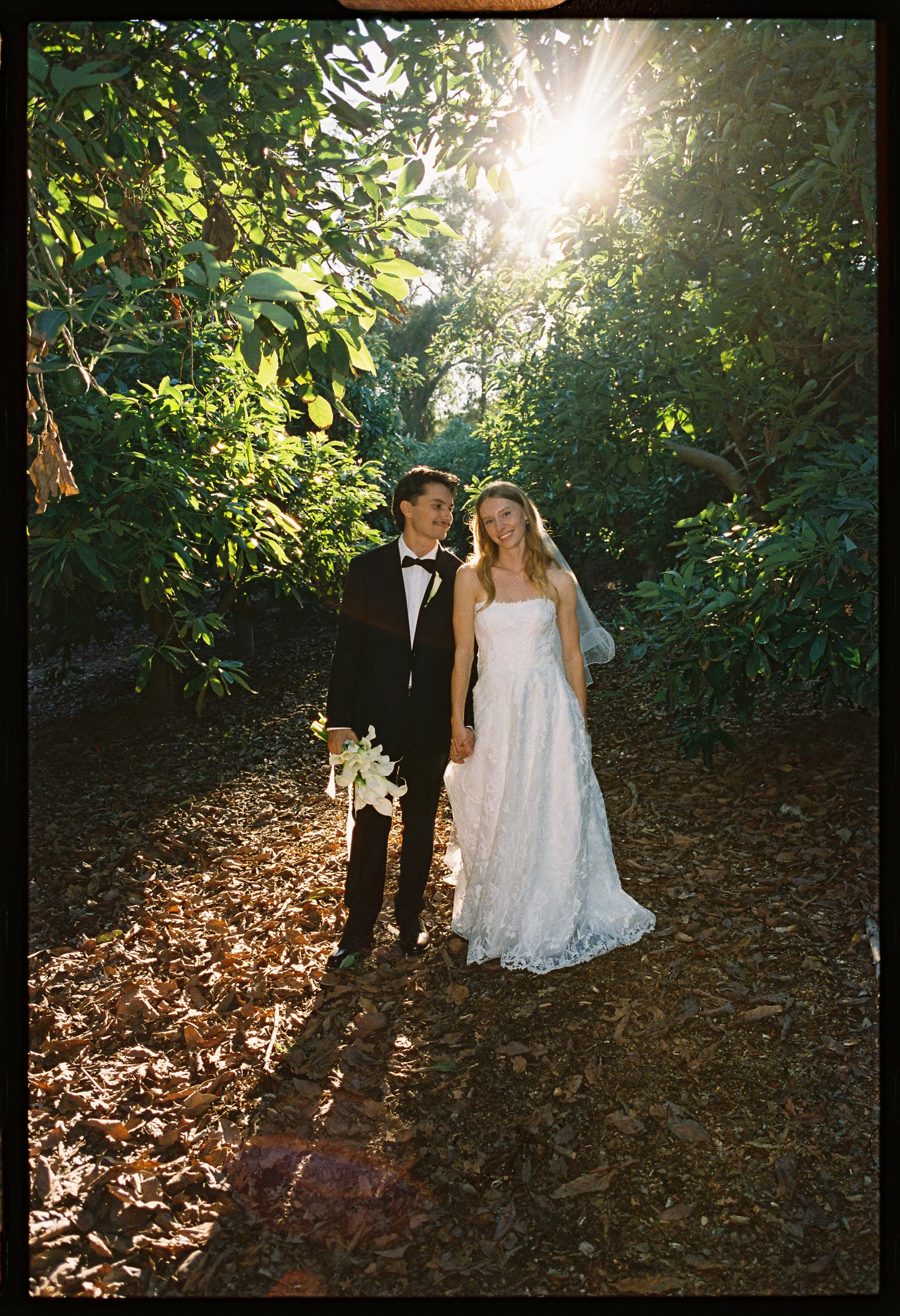 A bride and groom holding hands and walking on a wooded trail at sunset. The bride wears a white lace wedding dress and veil, holding a bouquet of white lilies. The groom wears a black tuxedo with a bow tie. Sunlight filters through the trees.