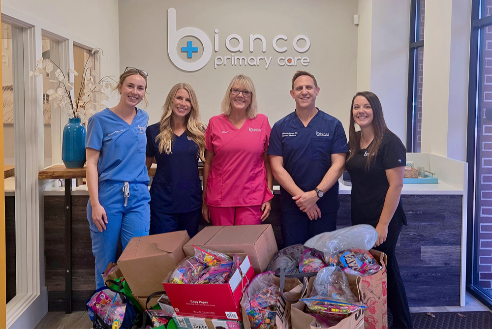 Group of five healthcare professionals standing behind multiple boxes of candy in a reception area for Bianco Primary Care, with a sign and window in the background.