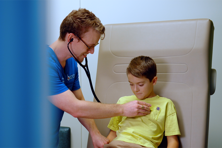 A male doctor using a stethoscope to examine a young boy in a hospital bed.