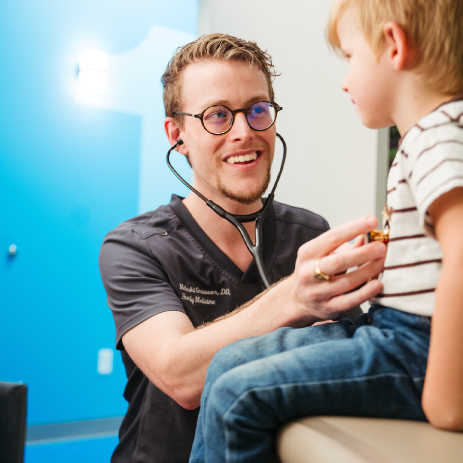 A male doctor with glasses and a stethoscope examining a young boy in a medical office.