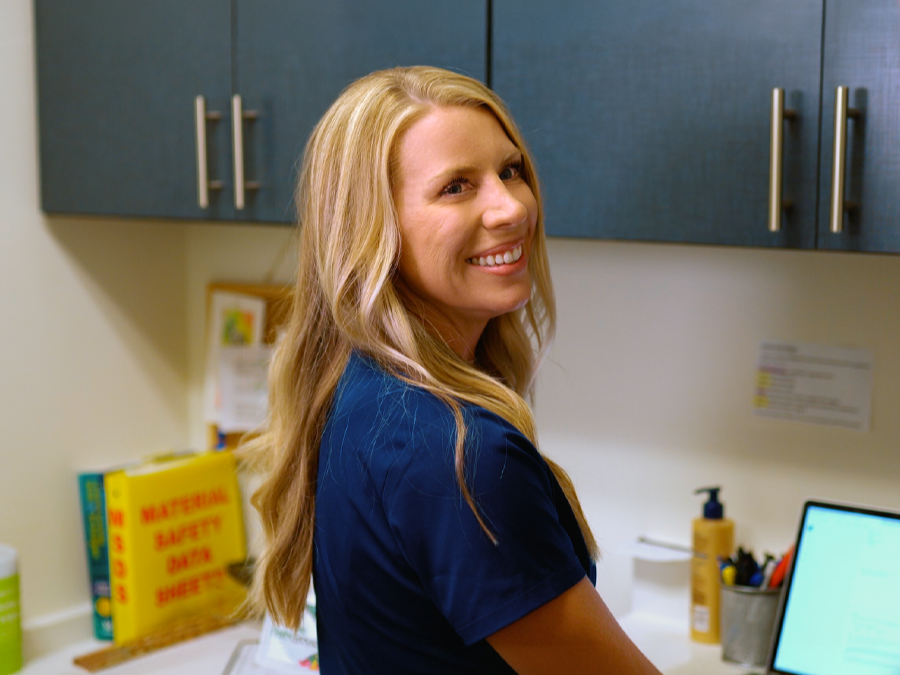 A woman with long blonde hair smiling at the camera in a medical or office setting.