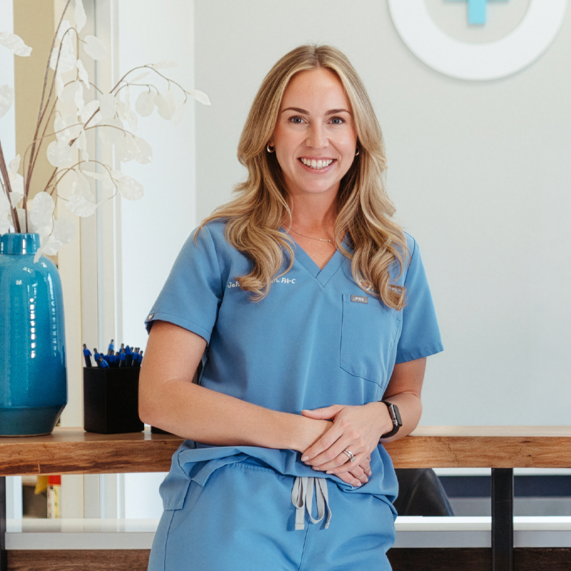 A smiling woman in blue medical scrubs leaning on a wooden reception counter in a healthcare setting.