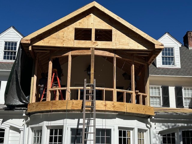 Under construction second-story balcony being built on a white house with multiple windows and a gray roof, using wooden framing and a ladder.