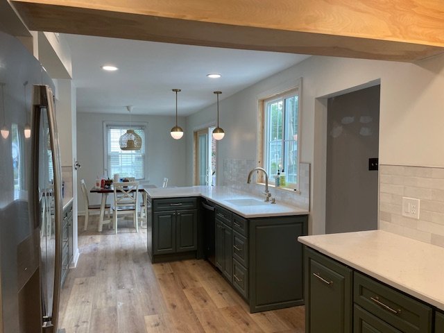 Modern kitchen with dark green cabinets, white marble countertops, stainless steel appliances, pendant lighting, and a dining area with a window in the background.