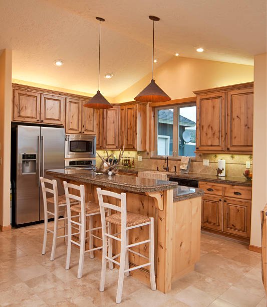 Kitchen with wooden cabinets, stainless steel refrigerator, built-in microwave, and an island with three white chairs. There are two hanging pendant lights above the island, a window above the sink, and warm lighting.