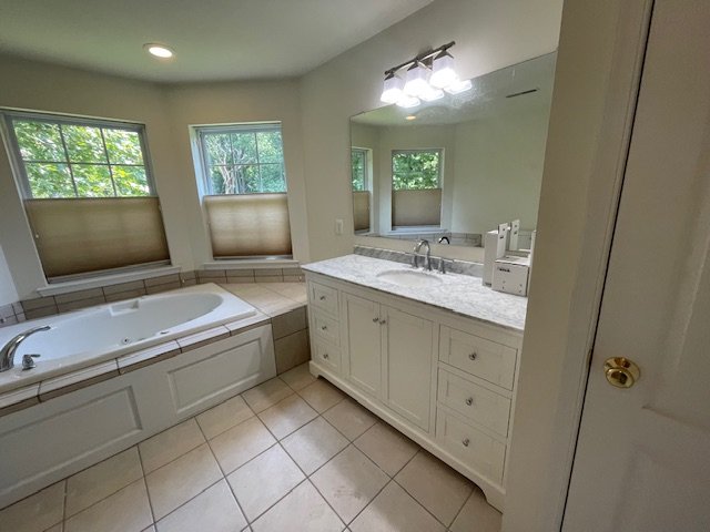 Bathroom with a large bathtub, a vanity with a sink, a mirror, and three windows with beige blinds.