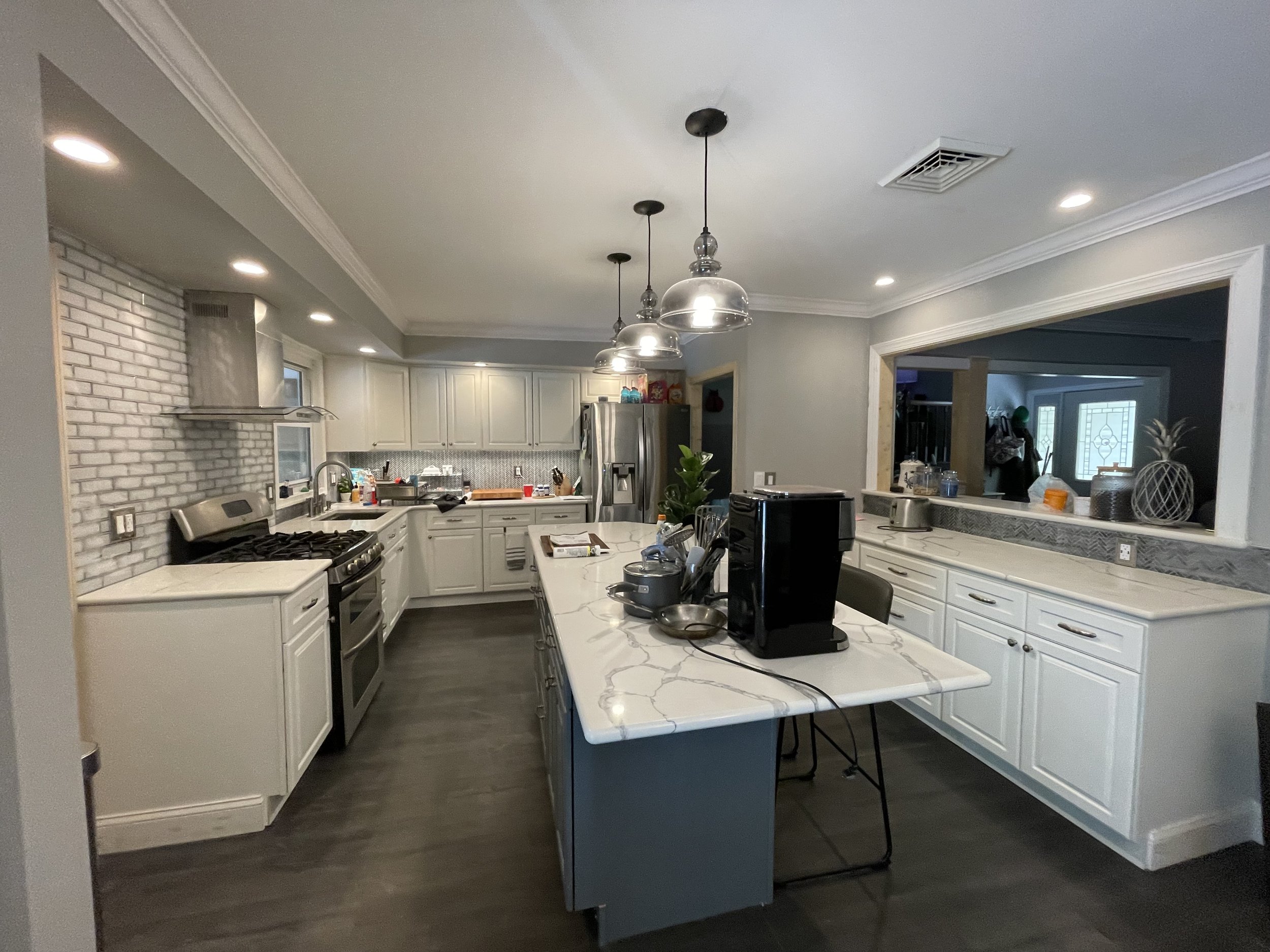 Modern kitchen with white cabinets, gray backsplash, and stainless steel appliances. Center island with marble countertop, pendant lights above, and a view into adjacent room.