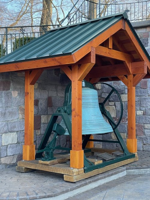 A large outdoor bell housed in a wooden shelter with a metal roof, placed on a wooden platform against a stone wall.