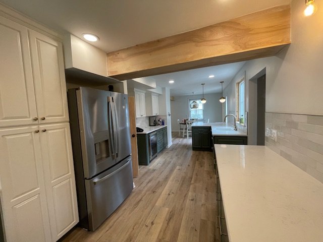 Modern kitchen with white cabinets, stainless steel refrigerator, wooden flooring, and a breakfast bar. In the background, there is a dining area with a table and chairs.