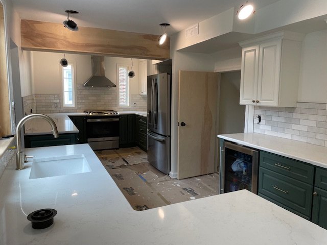 Kitchen with white countertops, green lower cabinets, white upper cabinets, stainless steel fridge, range, and hood, brick-style white tile backsplash, and a partially finished wood accent above the stove.