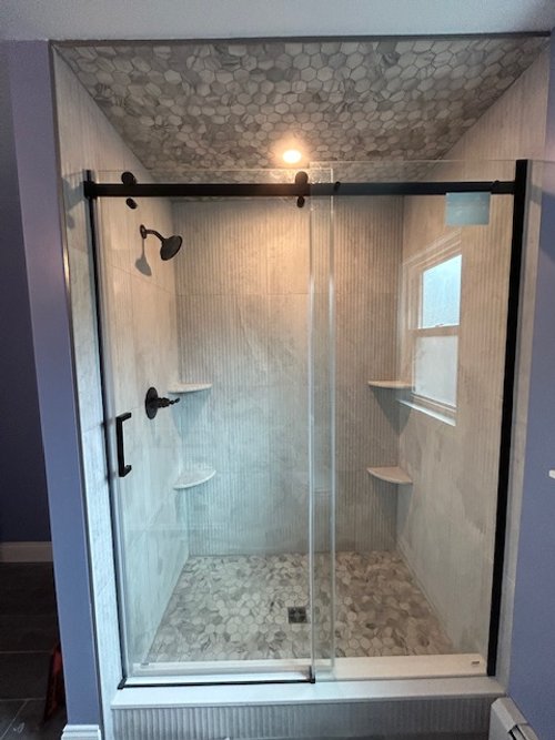 Inside a glass-enclosed walk-in shower with hexagonal tiles on the ceiling and pebble tiles on the floor, including a rainfall showerhead, a handheld showerhead, and built-in corner shelves.