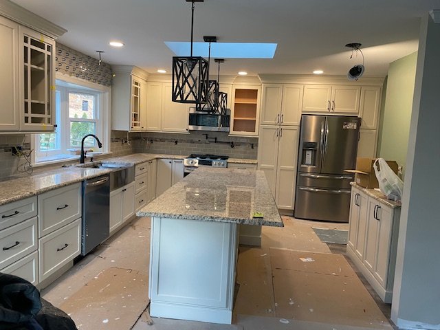 Kitchen with white cabinets, granite countertops, stainless steel refrigerator, island in center, pendant lighting, and ongoing installation work.